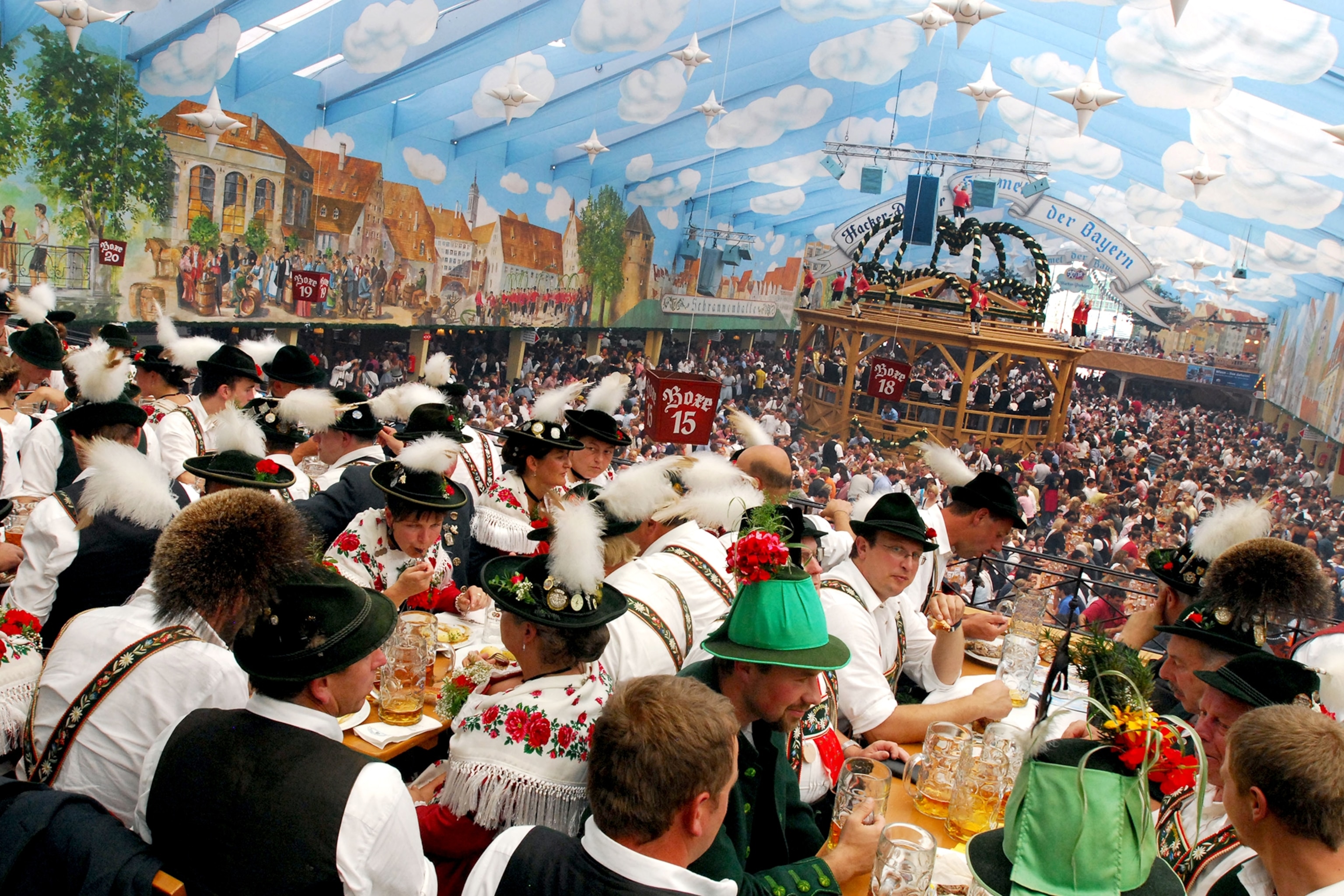 Revelers in tracht raising Maßkrüge at long tables inside the Hacker-Pschorr tent at Oktoberfest, Munich, under the tent's painted blue sky ceiling.