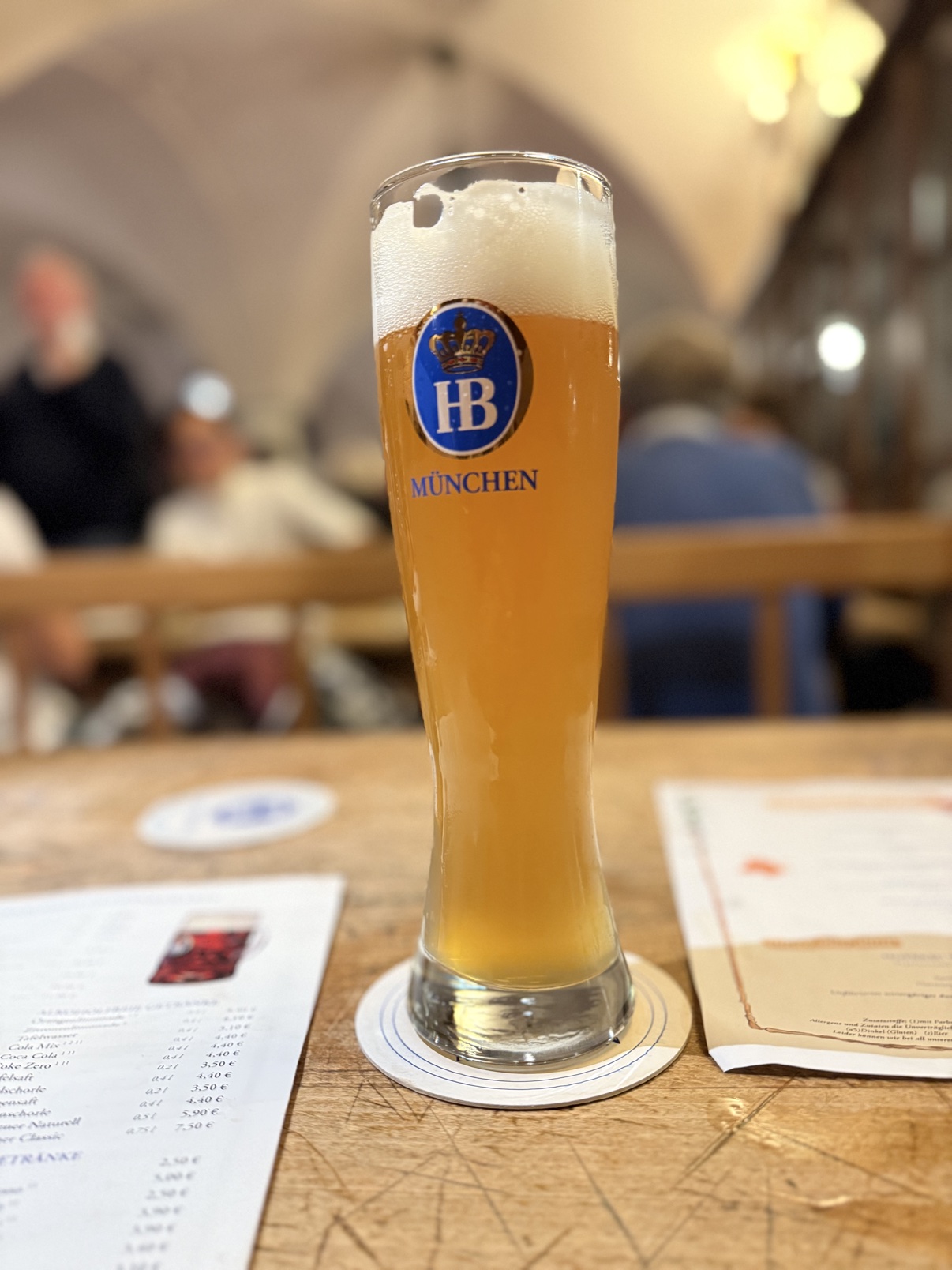 A tall Weizenglas of Hofbräu wheat beer with a thick, towering head, on a table inside the vaulted Hofbräuhaus beer hall.
