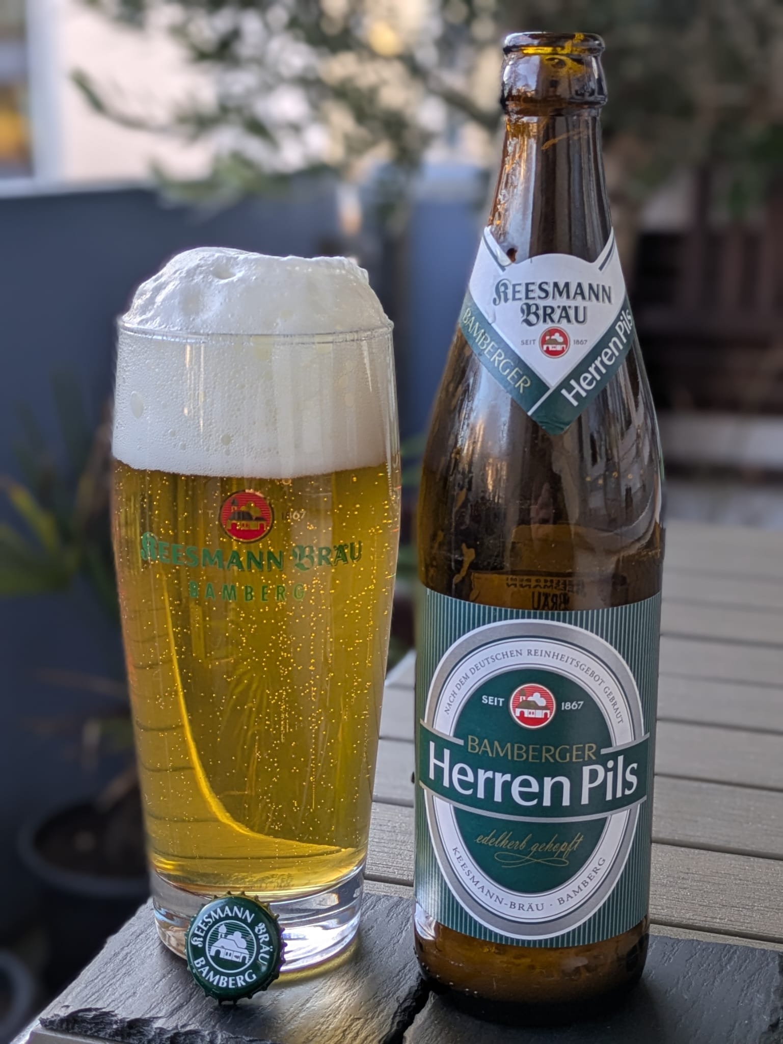 A poured glass of Keesmann Bamberger Herren Pils with a domed foam head, beside its bottle on a wooden table.
