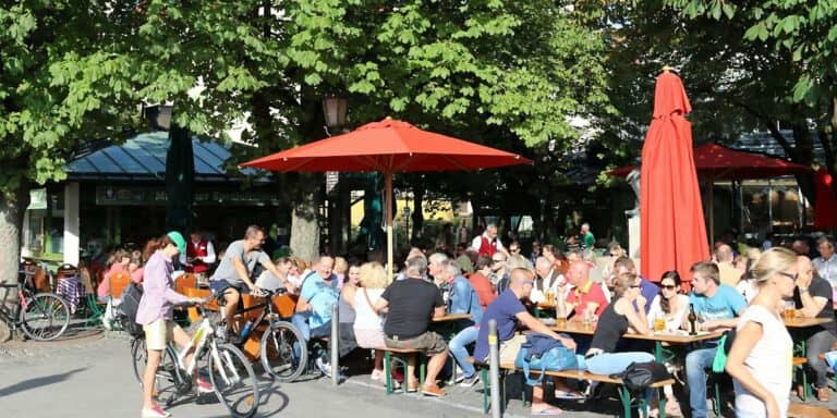 Guests seated at long tables under chestnut trees in the Biergarten Viktualienmarkt, in the middle of the Viktualienmarkt food market.