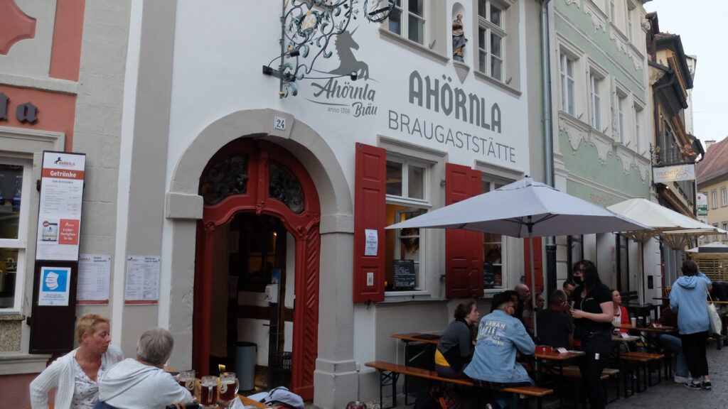 The Ahörnla Braugaststätte facade on Obere Sandstraße in Bamberg, with its wrought-iron horse sign, red shutters, and guests at outdoor tables.