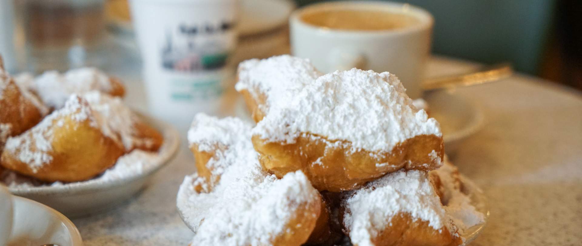 A plate of three sugar-dusted beignets piled high beside a steaming cup of café au lait on a speckled marble café counter, the classic New Orleans breakfast spread.