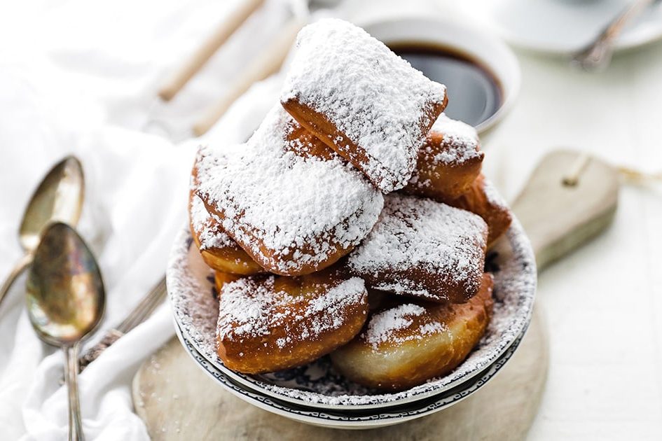 A plate of golden beignets dusted with powdered sugar next to a cup of milky café au lait on a marble counter at Morning Call's pavilion on Canal Boulevard in New Orleans.