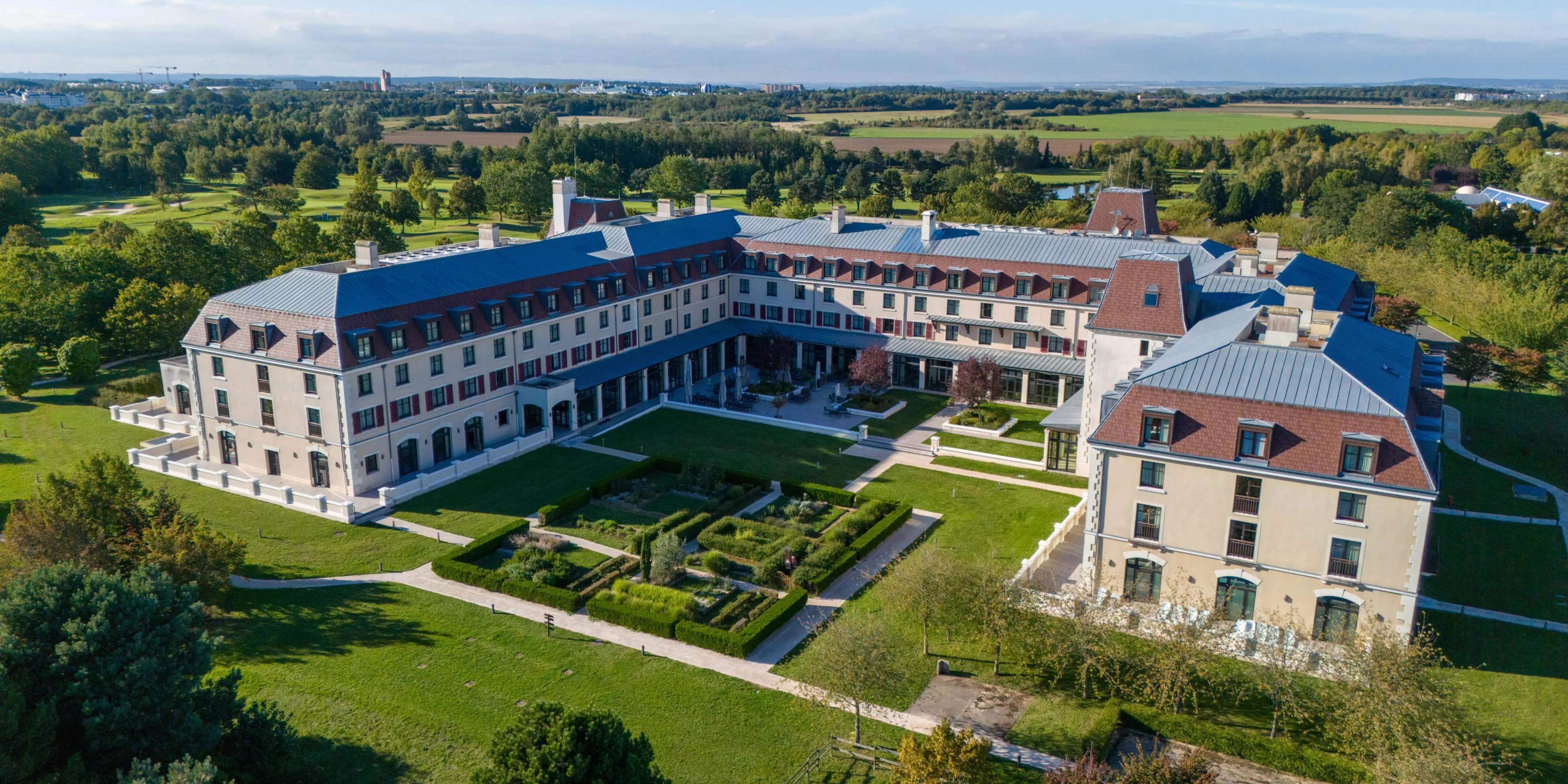 Aerial view of the Crowne Plaza Paris Marne-la-Vallée, courtyard buildings with red tile roofs set in landscaped grounds, golf course greens beyond.