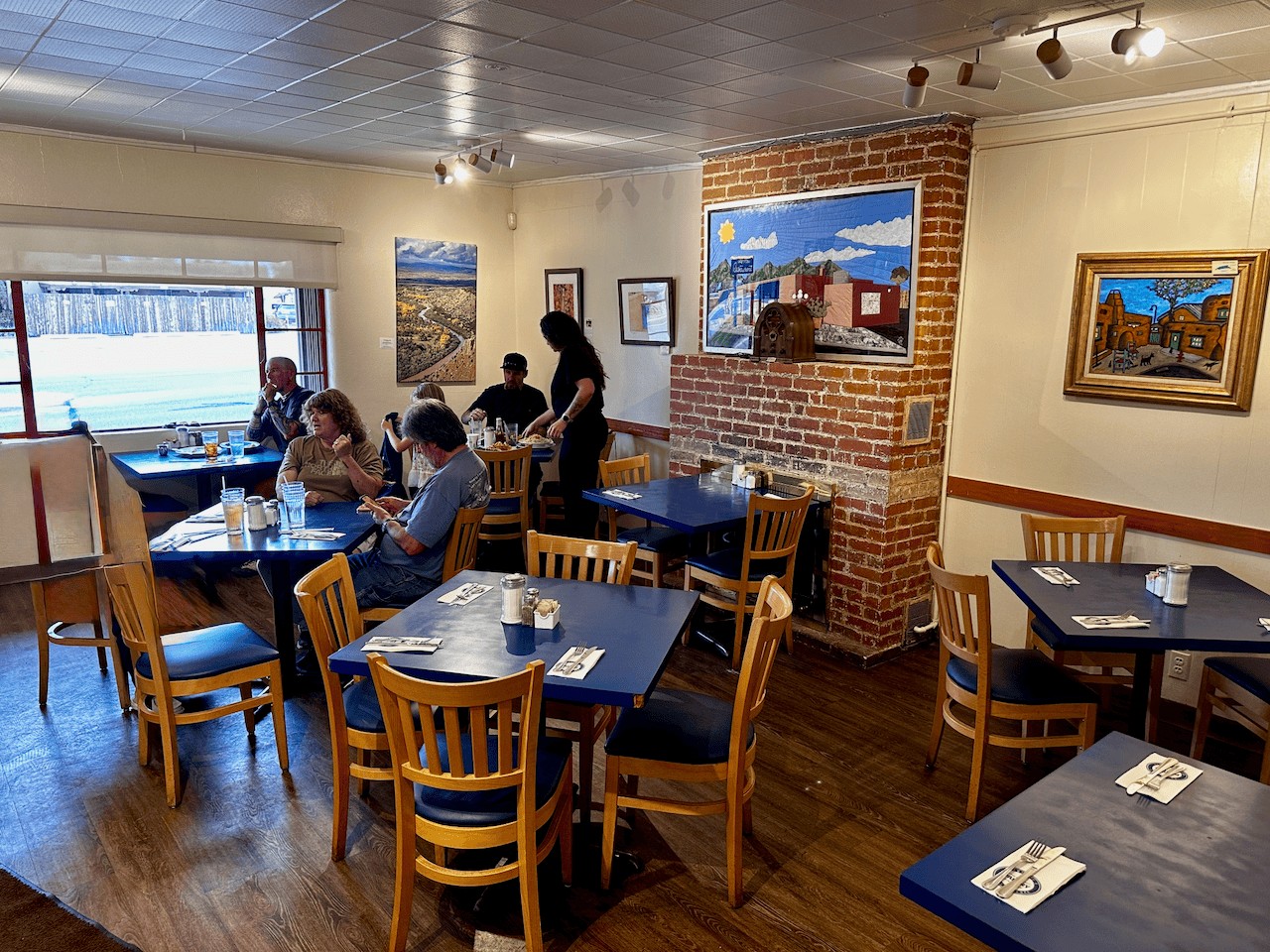 Inside The Pantry's front dining room in Santa Fe — blue tables, wood chairs, a brick fireplace with a quilt depicting the restaurant, and New Mexico art on the walls