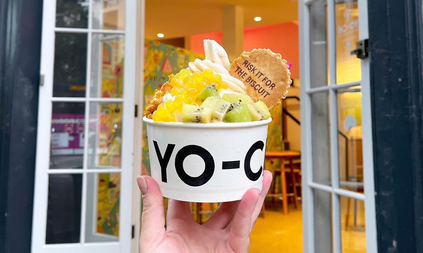 A loaded Yo-Chi branded cup with fro-yo swirl, popping pearls, kiwi, and a RISK IT FOR THE BISCUIT wafer, held up outside the Notting Hill store.